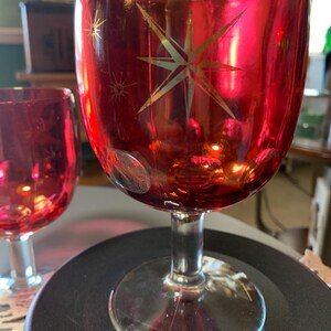May include: Two red wine glasses with gold starburst designs. The glasses have a clear stem and base. The red color is vibrant and the starbursts add a touch of elegance. Perfect for a holiday gathering or special occasion.