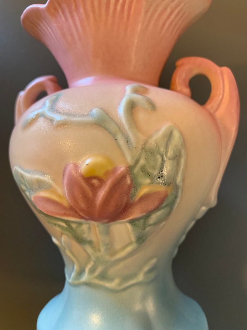 May include: A vintage ceramic vase with a pink, white, and blue glaze. The vase features a raised floral design with a pink flower and green leaves.