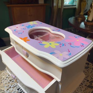 May include: A white wooden jewelry box with a lavender top featuring colorful floral patterns and a heart-shaped viewing window. The box includes a pull-out drawer with a small heart-shaped knob and an interior lined with soft pink material.