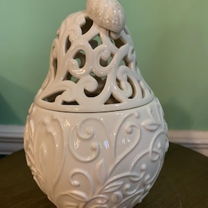 May include: A white pear-shaped decorative container with a lid. The container has an openwork design on the top half and embossed floral patterns on the bottom half. A small, textured pear-shaped accent sits on top.