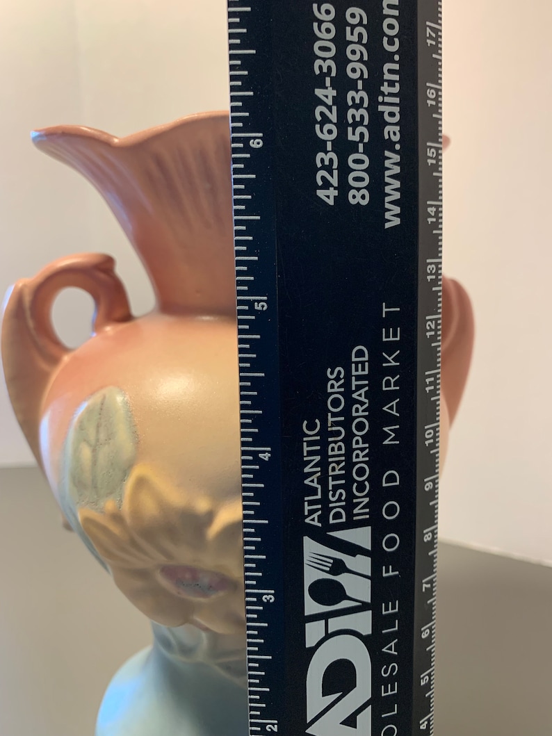 May include: A pink and white ceramic vase with a floral design. The vase is approximately 6 inches tall and has a wide mouth. The vase is sitting on a table next to a ruler.
