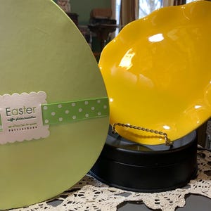 May include: A light green egg-shaped box with the word "Easter" and a green and white polka dot ribbon. A bright yellow ruffled plate sits on a black stand. The plate has a gold-colored handle.