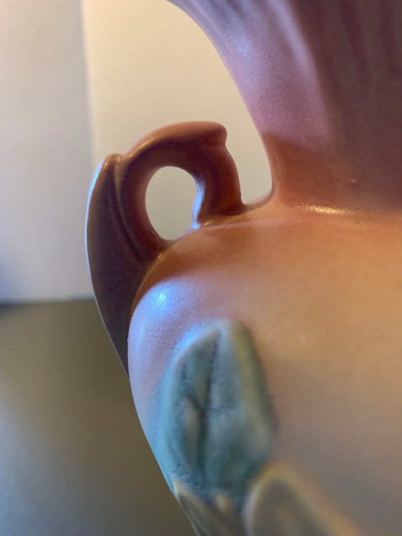 May include: A pink ceramic vase with a handle and a floral design. The vase has a textured surface and a glossy finish.