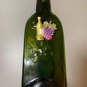 May include: A flattened, dark green glass bottle-shaped ornament. It features a gold-colored metal loop at the top for hanging. A small, detailed relief of a wine bottle and a bunch of grapes is affixed to the front. The grapes are purple and pink.
