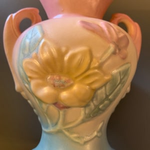 May include: A vintage ceramic vase with a pale pink top, a cream body, and a light blue base. The vase features a floral design with a large yellow flower, pink flowers, and green leaves.