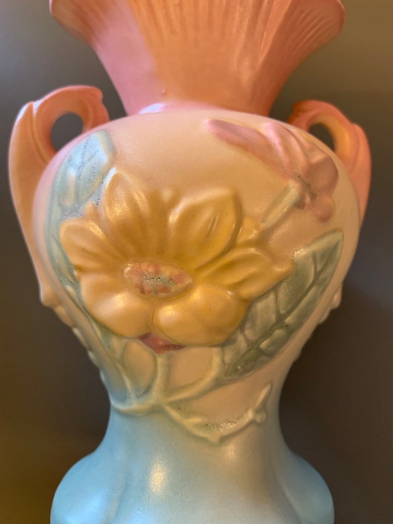 May include: A vintage ceramic vase with a pale pink top, a cream body, and a light blue base. The vase features a floral design with a large yellow flower, pink flowers, and green leaves.