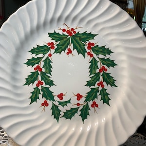 May include: White ceramic plate with a wavy rim design. The plate features a green holly wreath with red berries in the center. A gold-colored metal stand supports the plate, suggesting a decorative or serving piece for the holiday season.
