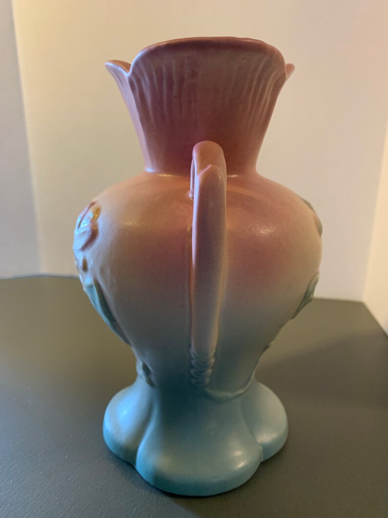 May include: A pink and blue ceramic vase with a handle and floral design. The vase has a gradient color scheme, transitioning from pink at the top to blue at the bottom.