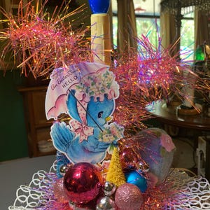 May include: A decorative Easter centerpiece featuring a central blue-tipped candle surrounded by iridescent pink tinsel. A vintage-style cutout of a blue bird holding an umbrella with the text An Easter Time HELLO is attached, accented by pink, silver, and blue ornaments.