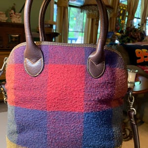 May include: A colorful handbag with a plaid pattern in shades of red, purple, blue, and brown. The bag has brown leather handles and accents, with a matching shoulder strap. The bag is a dome shape and has a zipper closure.