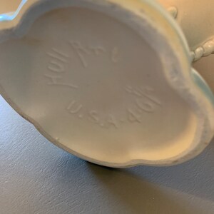 May include: The bottom of a white ceramic vase with a blue glaze. The vase has a scalloped edge and the words "Hallmark" and "U.S.A. 401" are printed on the bottom.