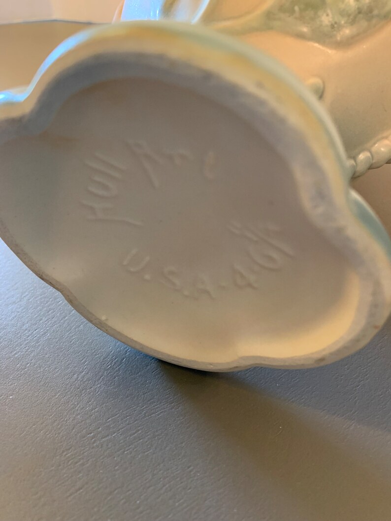 May include: The bottom of a white ceramic vase with a blue glaze. The vase has a scalloped edge and the words "Hallmark" and "U.S.A. 401" are printed on the bottom.
