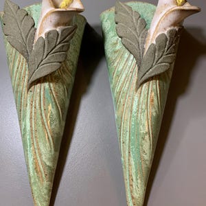 May include: A pair of cone-shaped ceramic wall vases featuring a textured light green finish with subtle brown accents. Each vase is adorned with a sculpted gray leaf overlay and a single white calla lily flower with a bright yellow spadix.