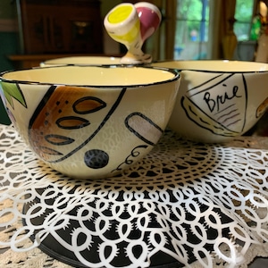 May include: A ceramic serving dish featuring three connected cream-colored bowls with hand-painted illustrations of food items, including a wedge of Brie cheese labeled with the text Brie, and a central handle topped with two small egg-shaped accents.