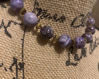 May include: A purple and white agate stone necklace with gold accents. The necklace is on a brown burlap background.