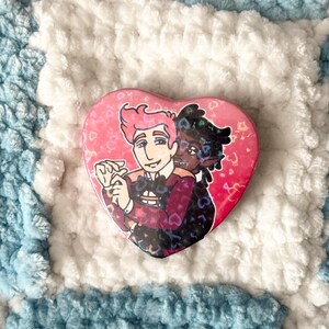 May include: A pink heart-shaped button featuring a cartoon illustration of two people holding hands. The people are dressed in formal attire and have different skin tones. The background is pink with white hearts.