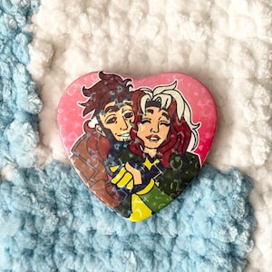 May include: A heart-shaped button with a cartoon image of two people hugging. The woman has long, white hair and is wearing a green and yellow outfit. The man has brown hair and is wearing a brown jacket. The background is pink.