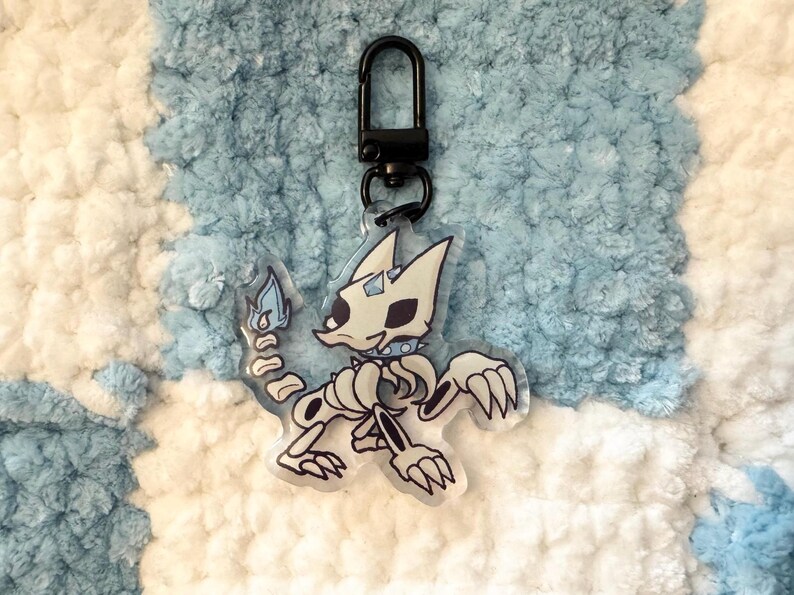 May include: A white and blue skeleton cat keychain with a black metal ring. The cat is holding a blue flame in its right paw.