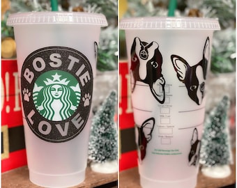 May include: A clear plastic reusable cup with a black and white design. The design features a black paw print with the words "Bostie Love" and a green Starbucks logo. The cup also has a black and white illustration of a French Bulldog on the side.