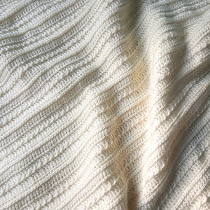 May include: A close-up of a white crocheted blanket with a textured pattern. The blanket is folded and draped, showing the soft, fluffy texture.