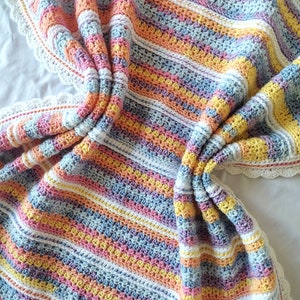Op de afbeelding: Een kleurrijk gestreept gehaakt babydekentje met een witte gehaakte rand. Het dekentje is gemaakt van verschillende pastelkleuren, waaronder roze, blauw, geel en oranje.