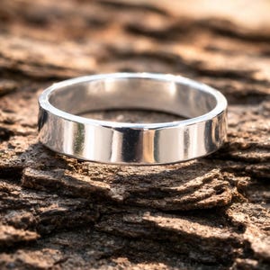 Könnte beinhalten: Ein silberner Bandring mit flacher, polierter Oberfläche. Der Ring befindet sich vor einem strukturierten, braunen Hintergrund mit verschwommenen grünen und gelben Tönen im Hintergrund.