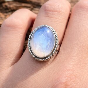Könnte beinhalten: Ein silberner Ring mit einem ovalen, schillernden Mondstein. Der Mondstein hat einen blau-weißen Farbverlauf. Der Ring hat ein gedrehtes Seildesign um den Stein. Der Ring befindet sich an einem Finger.