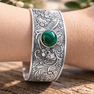 Könnte beinhalten: Ein silbernes Armreif-Armband mit aufwendigen floralen Mustern und einem ovalen Malachitstein in der Mitte. Das Armband ist breit und umschließt das Handgelenk, wobei detaillierte Gravuren zur Geltung kommen. Der Malachitstein ist ein leuchtendes Grün.