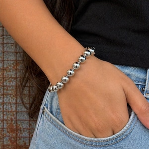 Op de afbeelding: Een zilveren kralenarmband met grote, ronde kralen. De armband wordt om een pols gedragen, met de hand in een spijkerbroekzak. De armband is gemaakt van glanzende, metalen kralen.