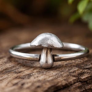 Könnte beinhalten: Ein silberfarbener Ring mit einem Pilz-Design. Der Pilz hat einen abgerundeten Hut und einen Stiel, der auf einem dünnen, kreisförmigen Band angebracht ist. Der Ring wird auf einer Holzoberfläche mit einem verschwommenen Hintergrund aus grünem Laub gezeigt.