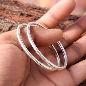 Könnte beinhalten: Zwei glänzende, silberne Armreifen werden in einer Hand gehalten. Die kreisförmigen Armreifen reflektieren das Licht. Der Hintergrund ist eine verschwommene Naturkulisse mit Braun- und Rosatönen.