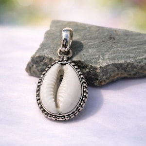 Könnte beinhalten: Silberanhänger mit einer weißen Kaurischnecke. Die Muschel ist in einem Silberrahmen mit einem Perlenrand und einer Schlaufe für eine Kette gefasst. Der Anhänger wird auf einem grauen Stein vor einem weichen, verschwommenen Hintergrund gezeigt.