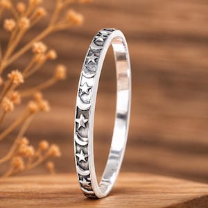Könnte beinhalten: Ein silberfarbener Metallring mit einem sich wiederholenden Muster aus geprägten Sternen und Mondsicheln auf dem äußeren Band. Das Design weist ein abgedunkeltes, oxidiertes Finish in den vertieften Bereichen auf, um die Motive hervorzuheben.