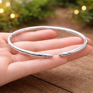 Könnte beinhalten: Ein silberfarbenes Armreif-Armband mit minimalistischem Design. Das offene Armband hat kugelartige Spitzen. Das glatte, polierte Metall reflektiert das Licht und erzeugt ein helles, glänzendes Aussehen. Dieses Schmuckstück ist für den täglichen Gebrauch geeignet.