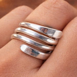 Könnte beinhalten: Ein silberfarbener Ring mit Spiraldesign. Der Ring umschlingt den Finger mit vier Bändern und erzeugt eine moderne, minimalistische Ästhetik. Das Metall hat eine polierte, reflektierende Oberfläche.