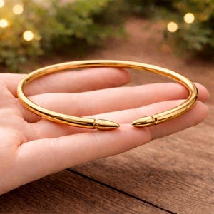 Könnte beinhalten: Ein goldfarbenes, offenes Armreif-Armband. Das Armband hat eine glatte, polierte Oberfläche und spitze, kugelartige Enden. Der Schmuck wird auf einer Hand präsentiert, mit einem verschwommenen Hintergrund aus Grün und Lichtern.