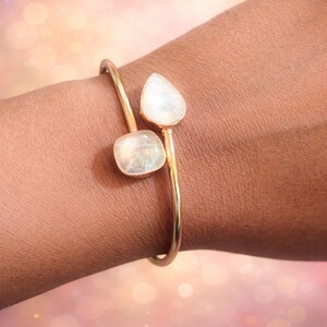 Könnte beinhalten: Ein goldfarbenes Armreif-Armband mit zwei schillernden, rechteckigen und tropfenförmigen Steinen. Das Armband umschließt das Handgelenk und betont die subtilen Farben der Edelsteine. Das Armband ist ein Schmuckstück.