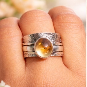 Può includere: Un anello d'argento con una pietra preziosa ovale color ambra. L'anello ha un motivo floreale sulla fascia e un centro rotante. La pietra è incastonata in una lunetta d'argento.