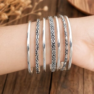 Könnte beinhalten: Nahaufnahme einer Sammlung silberner Armreifen an einem Handgelenk. Die Armreifen haben unterschiedliche Designs, darunter glatte Bänder und Bänder mit floralen und geometrischen Mustern. Die Armbänder werden zusammen getragen, wodurch ein geschichteter Look entsteht. Schmuck.
