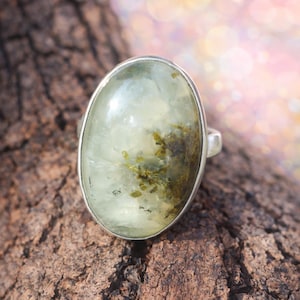 Könnte beinhalten: Ovaler Ring mit einem hellgrünen Edelstein mit dunkleren grünen Einschlüssen. Der Ring ist in einem silberfarbenen Metallband gefasst. Er liegt auf einer strukturierten, braunen Holzoberfläche. Der Stein ist etwa 2,5 cm lang.