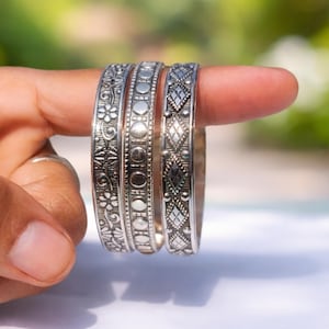 Könnte beinhalten: Drei silberne Armreifen mit aufwendigen Mustern werden gezeigt. Die Armreifen weisen florale, kreisförmige und rautenförmige Designs auf. Der Schmuck wird von einem Finger gehalten, wodurch die detaillierte Handwerkskunst und Textur des Metalls hervorgehoben werden.
