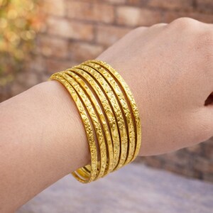 Könnte beinhalten: Eine Sammlung goldener Armreifen, jeder mit aufwendigen floralen Mustern verziert. Die Armreifen sind an einem Handgelenk gestapelt und zeigen ihr detailliertes Design und ihren strahlenden, goldenen Farbton. Der Schmuck wird vor einem verschwommenen Hintergrund präsentiert.