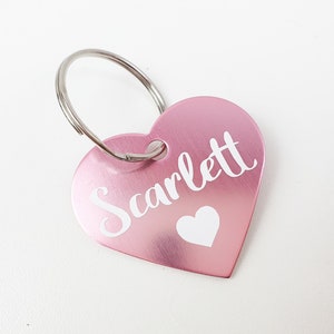 K&ouml;nnte beinhalten: Ein rosafarbener herzf&ouml;rmiger Schl&uuml;sselanh&auml;nger aus Metall mit dem Namen "Scarlett" in wei&szlig;er Schrift und einem kleinen wei&szlig;en Herz.