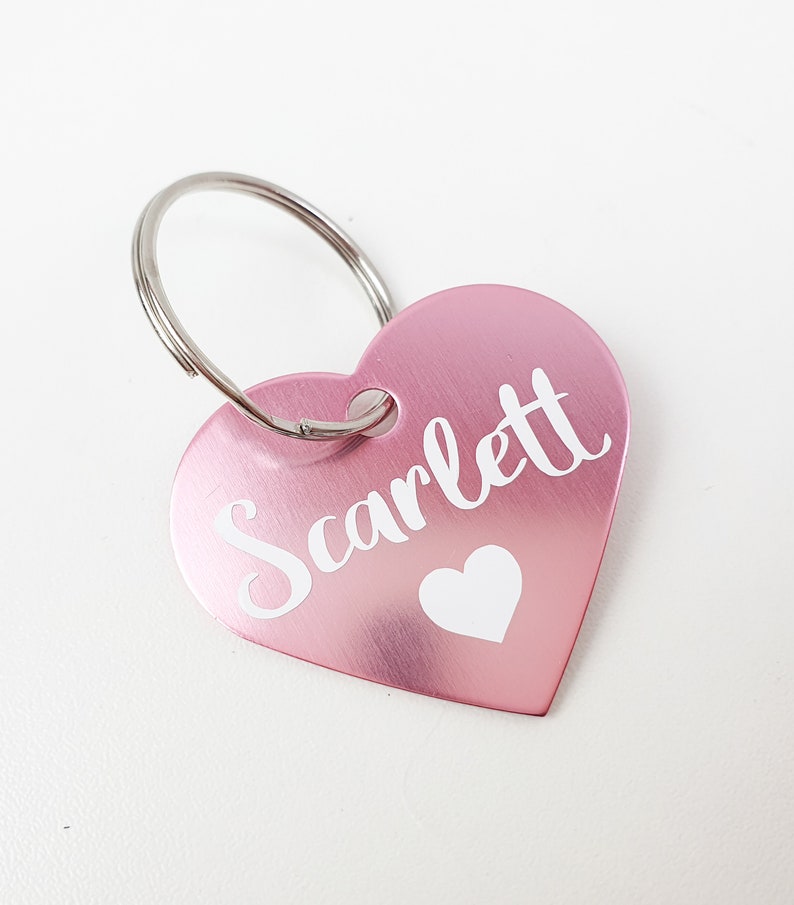 K&ouml;nnte beinhalten: Ein rosafarbener herzf&ouml;rmiger Schl&uuml;sselanh&auml;nger aus Metall mit dem Namen "Scarlett" in wei&szlig;er Schrift und einem kleinen wei&szlig;en Herz.