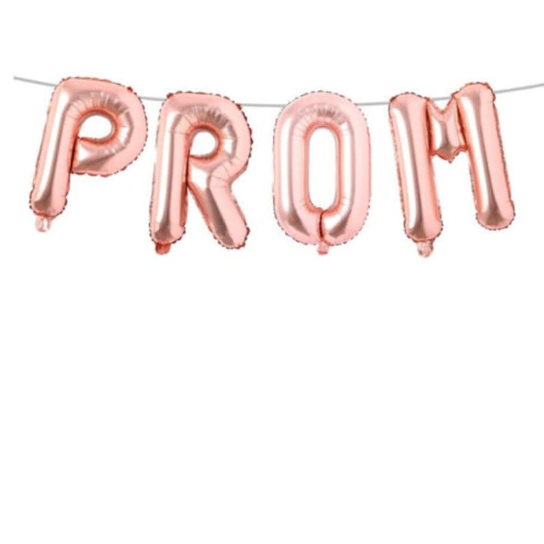 May include: Rose gold foil balloons spelling out the word "PROM" are strung on a thin, silver string. The balloons are inflated and have a metallic sheen, perfect for a prom celebration. The background is plain white.