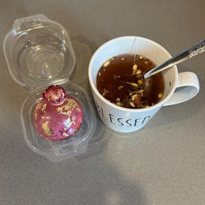 May include: A pink, spherical dessert with gold leaf accents sits in a clear plastic container. Beside it, a white mug with the word "BLESSED" contains tea with a tea bag and a spoon.