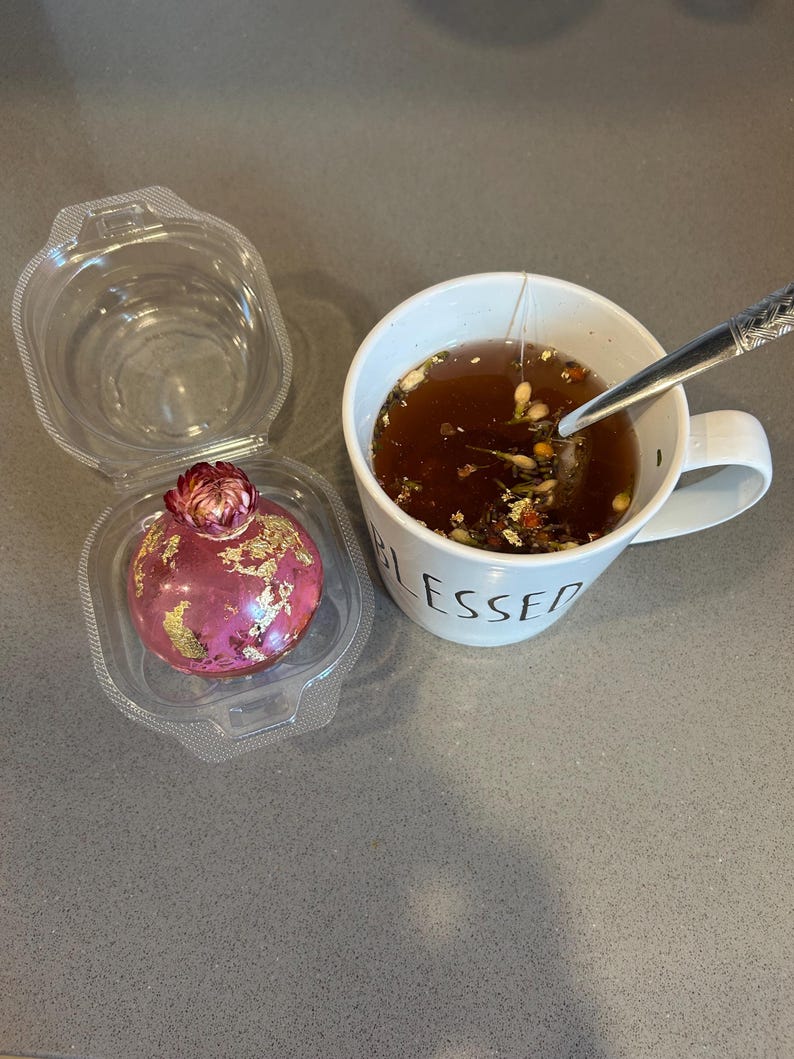 May include: A pink, spherical dessert with gold leaf accents sits in a clear plastic container. Beside it, a white mug with the word "BLESSED" contains tea with a tea bag and a spoon.