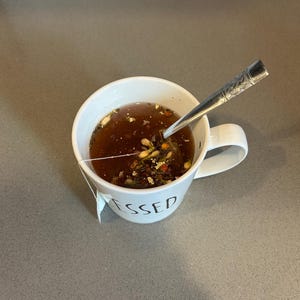 May include: A white ceramic mug filled with tea and a tea bag. The mug has the word "BLESSED" printed on it. A silver spoon rests inside the mug. The tea is a dark brown color and contains visible herbs and spices.