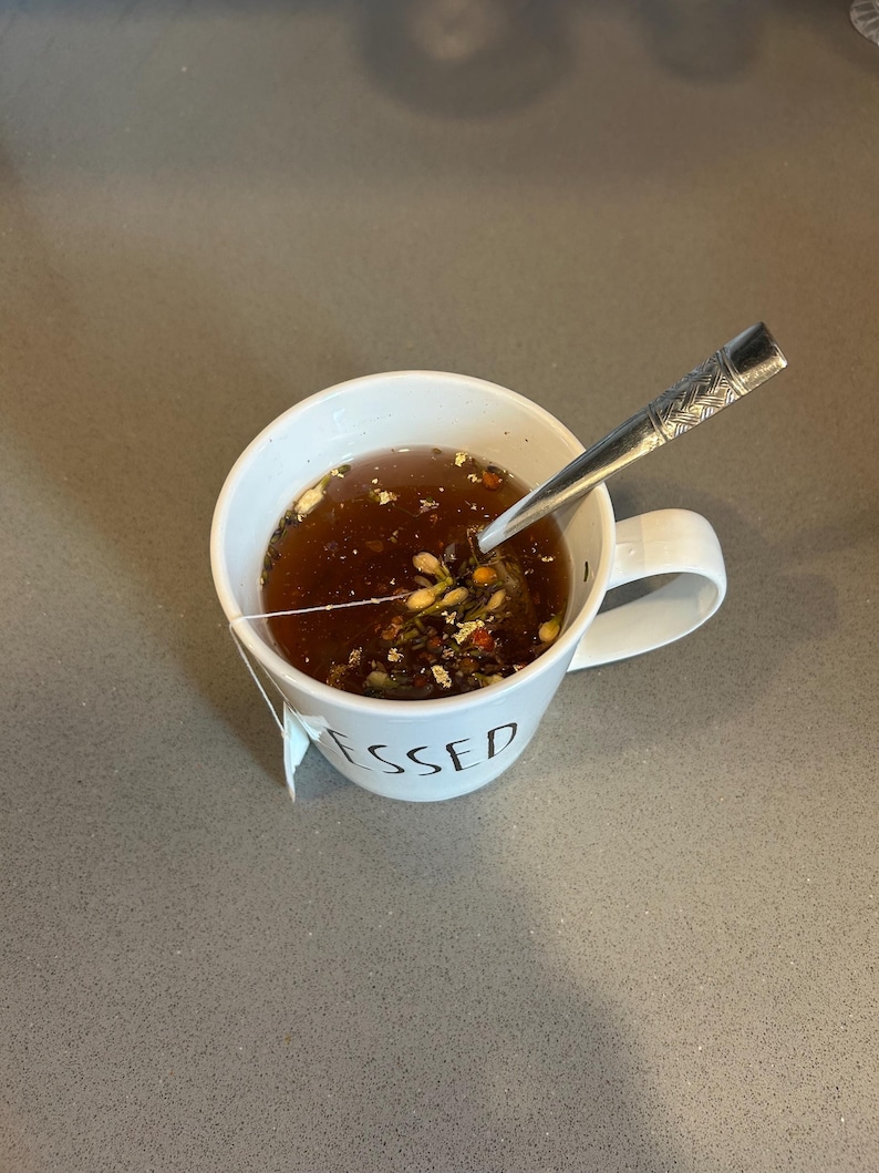 May include: A white ceramic mug filled with tea and a tea bag. The mug has the word "BLESSED" printed on it. A silver spoon rests inside the mug. The tea is a dark brown color and contains visible herbs and spices.