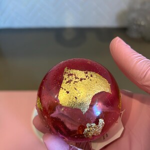 May include: A round, translucent, red object with gold leaf accents. The object is held in a gloved hand. The gold leaf is irregularly shaped and adds a decorative element to the red sphere.